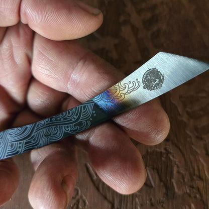 Hand holding a small knife with decorative blade and handle on a wooden surface