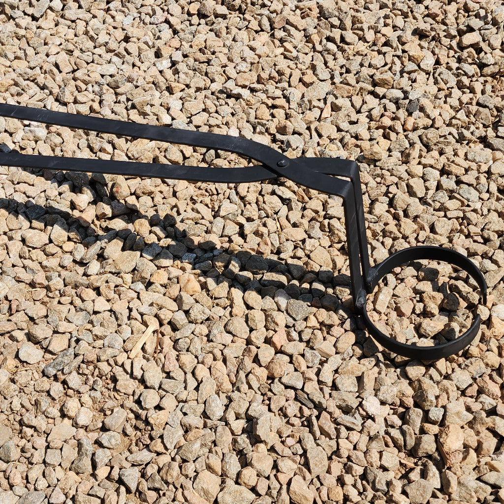 A set of black crucible tongs laid on a gravel surface.